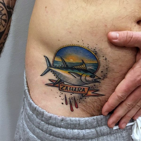 Traditional-style tuna fish tattoo on a man's hip, featuring vibrant colors, a sunset background, and a "Zahara" banner for a nautical theme.