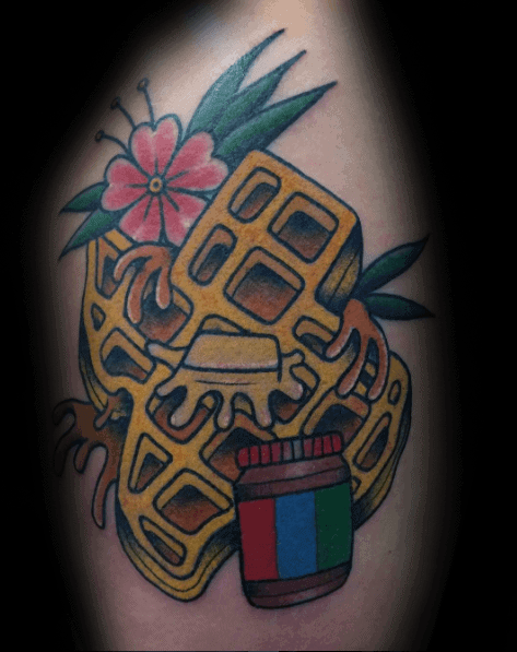 Traditional-style waffle tattoo with dripping syrup, a butter pat, a peanut butter jar, and tropical flowers, combining bold colors with a classic design.