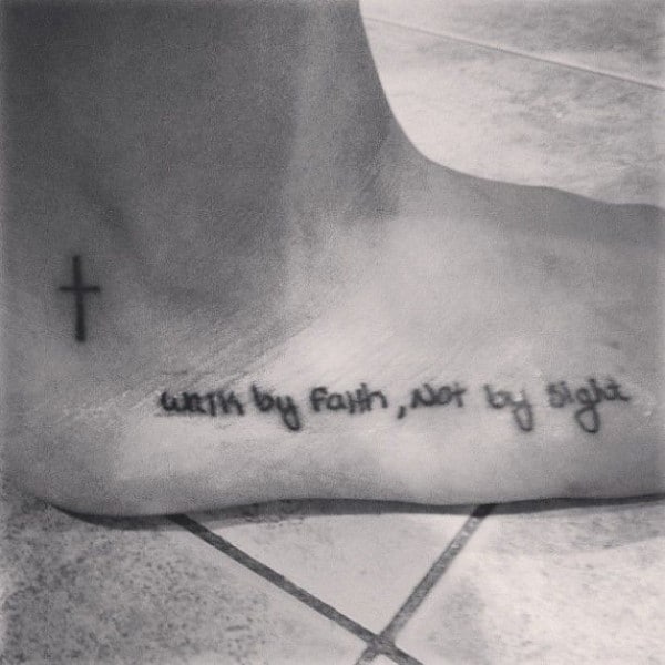 Minimalist ankle tattoo featuring a small cross and the phrase walk by faith, not by sight in delicate handwritten-style script.