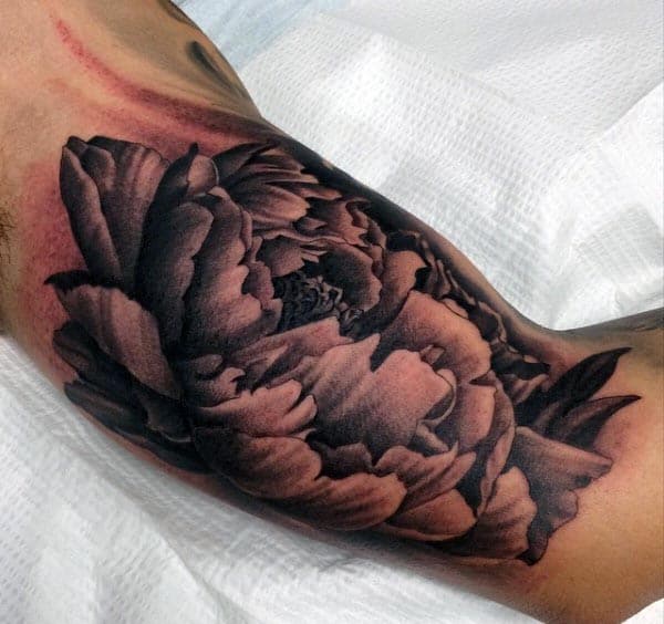 Black and grey tattoo of a large, detailed peony flower on the inner bicep, featuring soft shading and realistic petal textures
