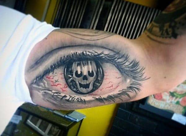 Black and grey tattoo of a realistic eye on the inner bicep, with a skull reflection in the pupil and red veins for a haunting effect