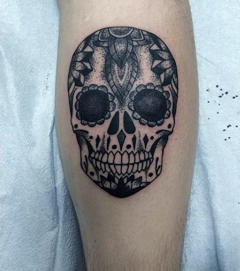 Black ink calf tattoo of a sugar skull with intricate floral and geometric patterns, inspired by Día de los Muertos designs