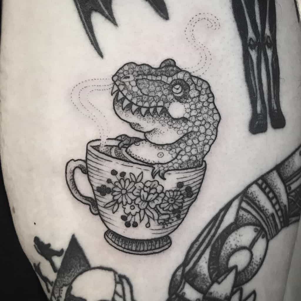 Funny tattoo of a tiny T-Rex soaking in a teacup, steam rising as if it's a relaxing hot bath. Detailed linework with floral design.