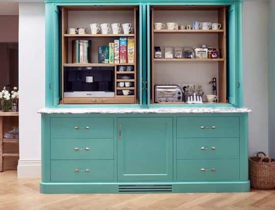 Teal coffee bar with marble countertop, open shelves, built-in coffee machine, and mugs.