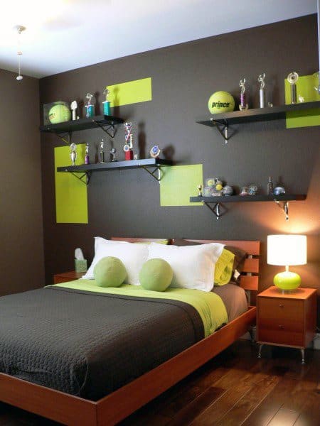 Modern bedroom with green accents, displaying trophies on shelves above the bed, wooden floor, and soft lighting