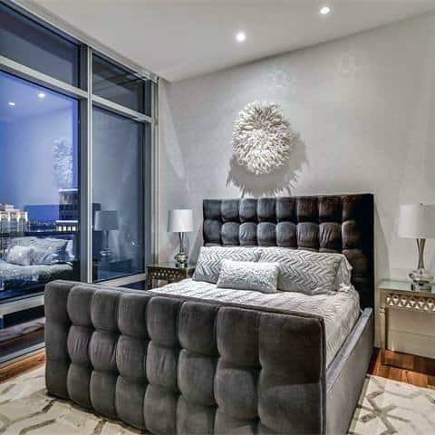 Modern bedroom with a large tufted bed, bedside tables, lamps, and a city view through floor-to-ceiling windows