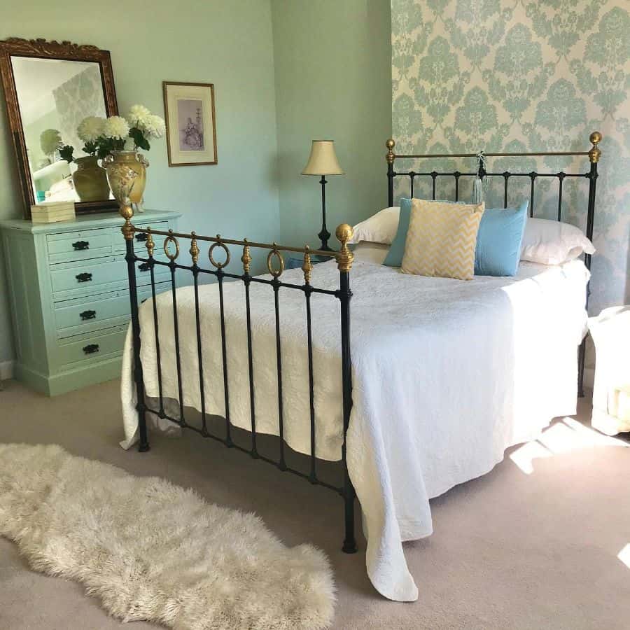 Vintage bedroom with an iron bed, pastel green walls, ornate dresser, and fluffy rug. Soft lighting fills the room