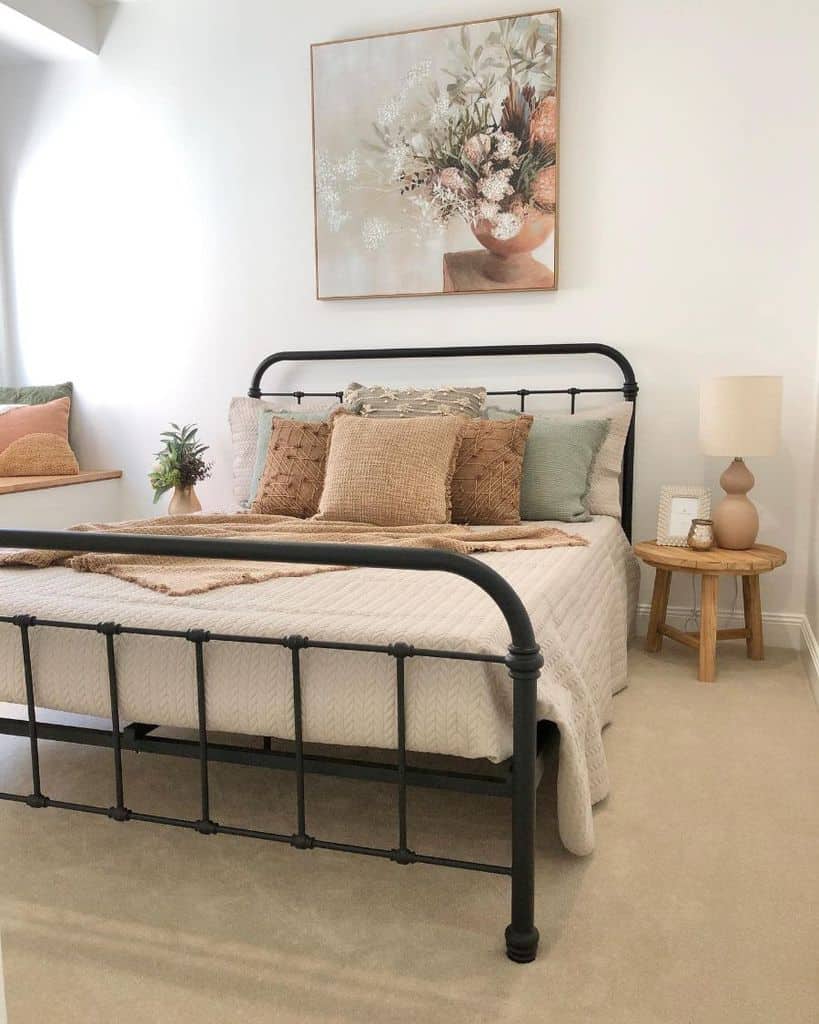 Cozy bedroom with a metal bed, pillows, a side table with a lamp, and a floral painting on the wall