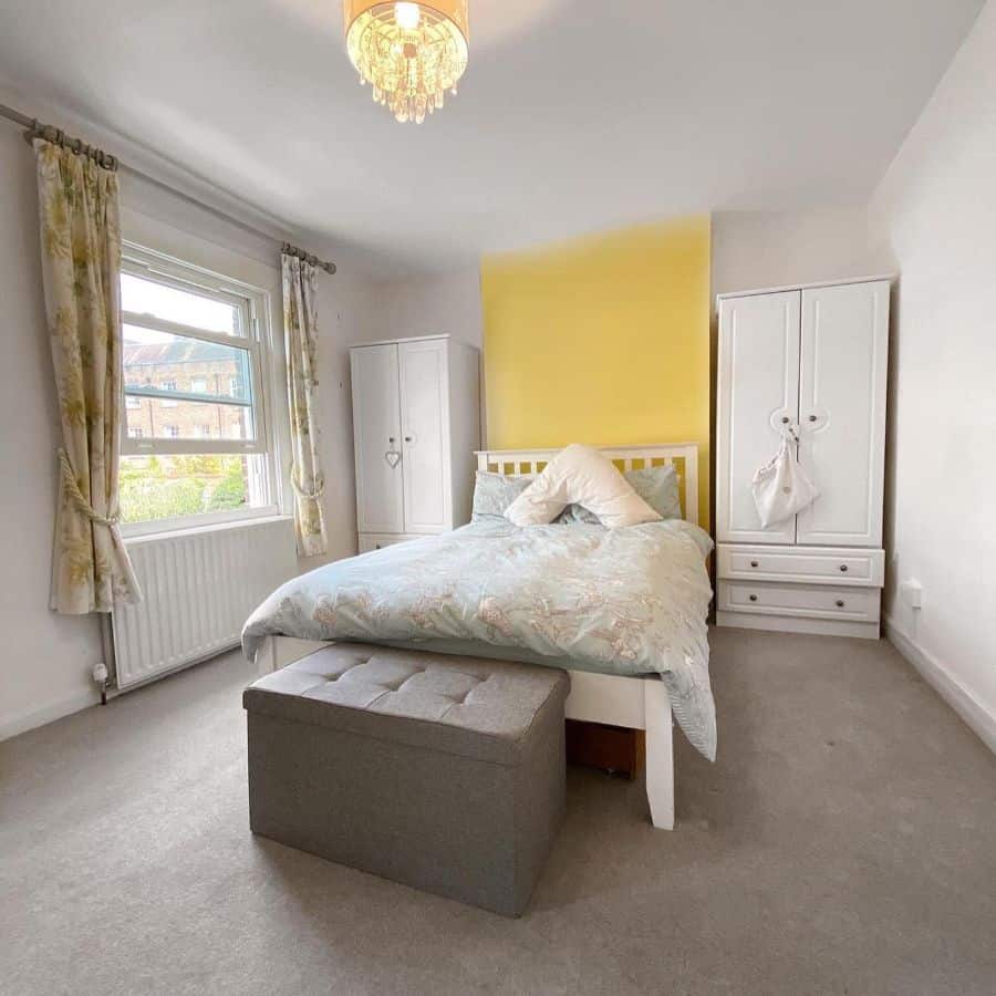 A bedroom featuring a yellow accent wall, a white bed, a gray bench, and two white wardrobes beside the bed