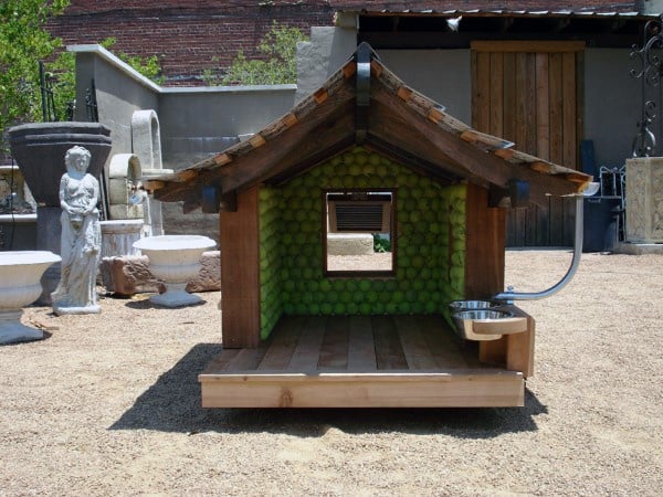 A unique doghouse with a green tennis ball interior and an attached stainless steel water bowl on a sandy yard