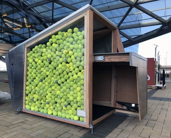 Dog house with a wall filled with green tennis balls, featuring a modern angular design, located in an open space near a building