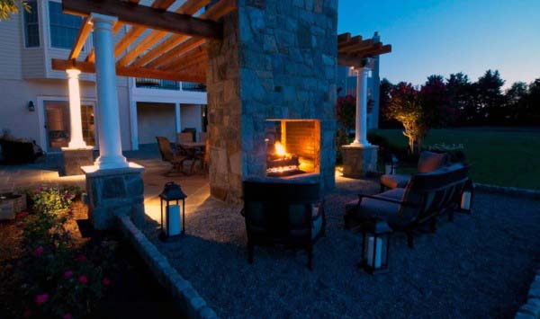 Outdoor patio with lit stone fireplace, cozy seating, lanterns, and pergola at dusk