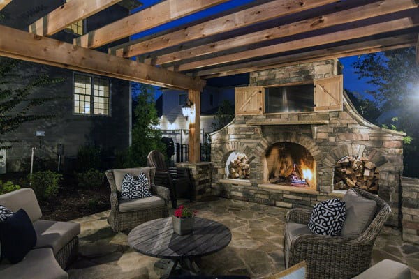 Cozy outdoor patio at night with stone fireplace, wooden pergola, and comfortable wicker furniture