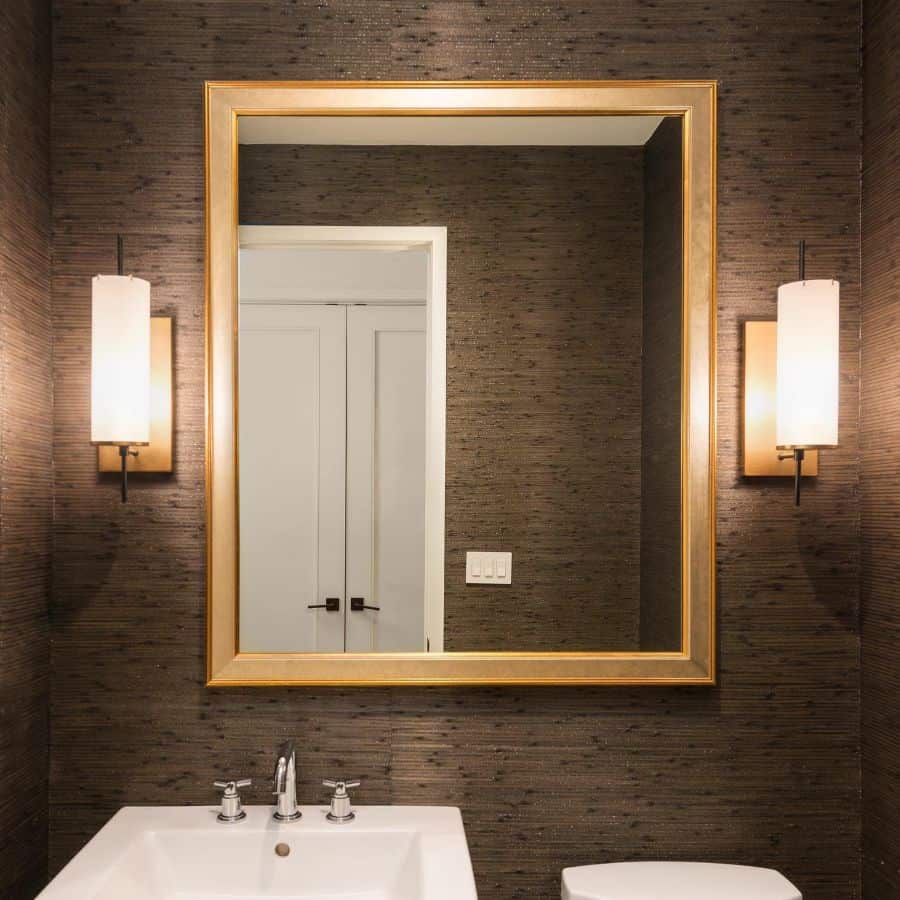 Modern bathroom with brown textured walls, gold-framed mirror, and wall sconces.