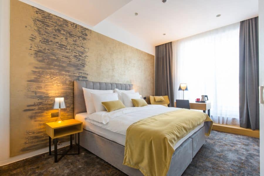 Cozy hotel room with a double bed, yellow bedding, an accent wall, bedside lamps, and a desk by a large window