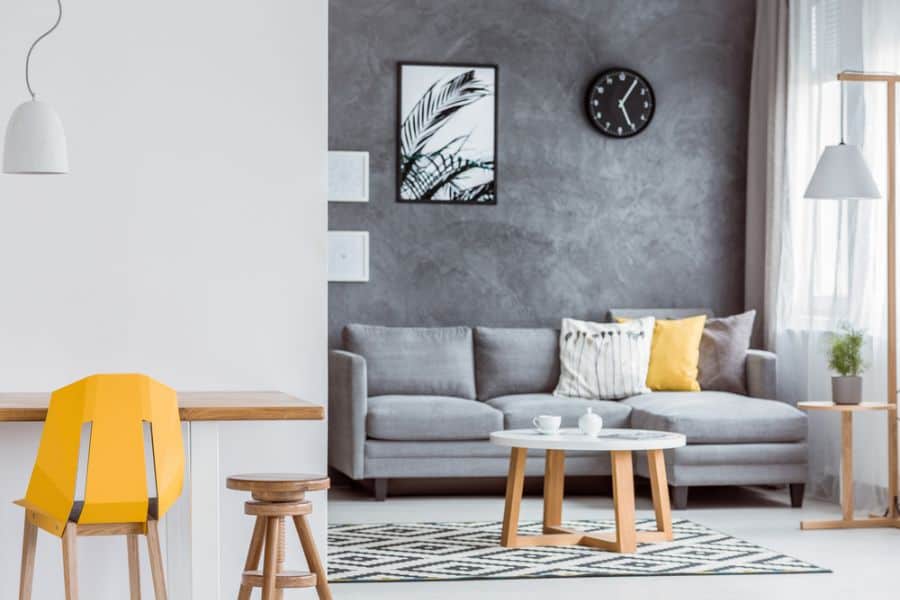 Modern living room with a grey sofa, yellow chair, wooden accents, and an accent wall featuring a black clock and plant print