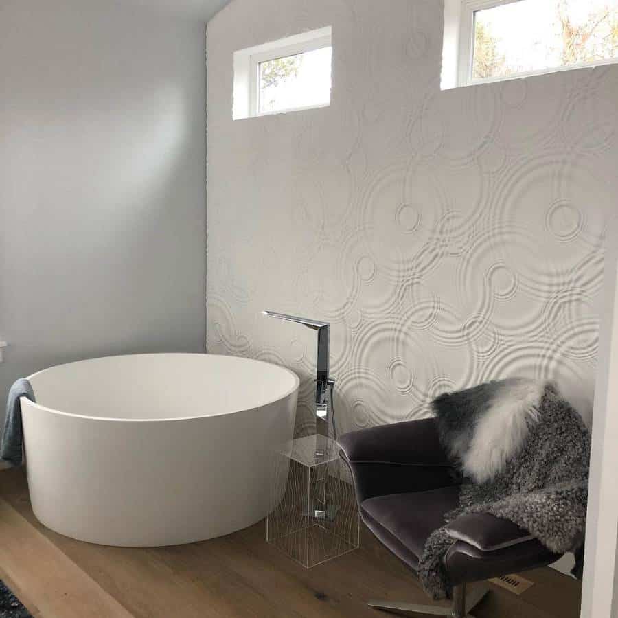 Modern bathroom with round tub, textured accent wall, dark chair with fur throw, and a small clear side table