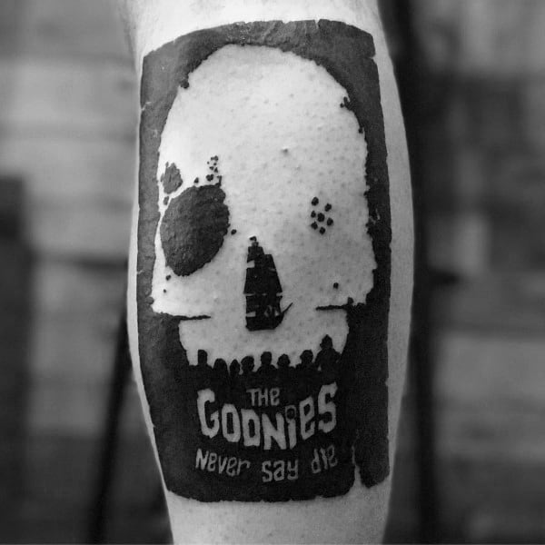 Black and gray minimalist tattoo of *The Goonies*, featuring a skull silhouette with a pirate ship inside and the phrase "The Goonies Never Say Die".