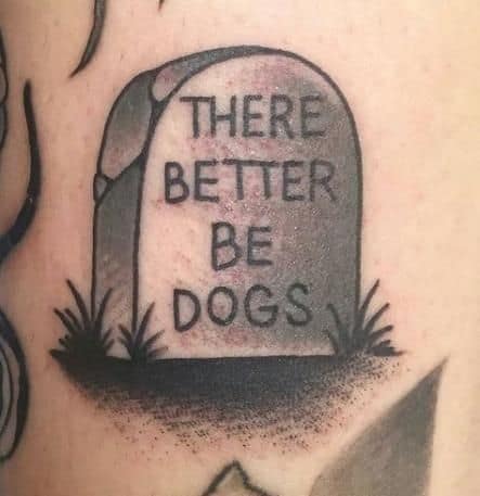 A black ink tattoo of a tombstone with the humorous inscription 'THERE BETTER BE DOGS,' shaded for a classic grave marker look.