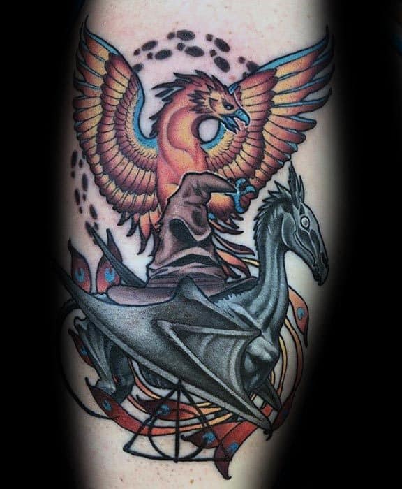 Colorful tattoo featuring a Thestral and a Phoenix with the Sorting Hat, dynamic composition and bold contrast on the upper arm.