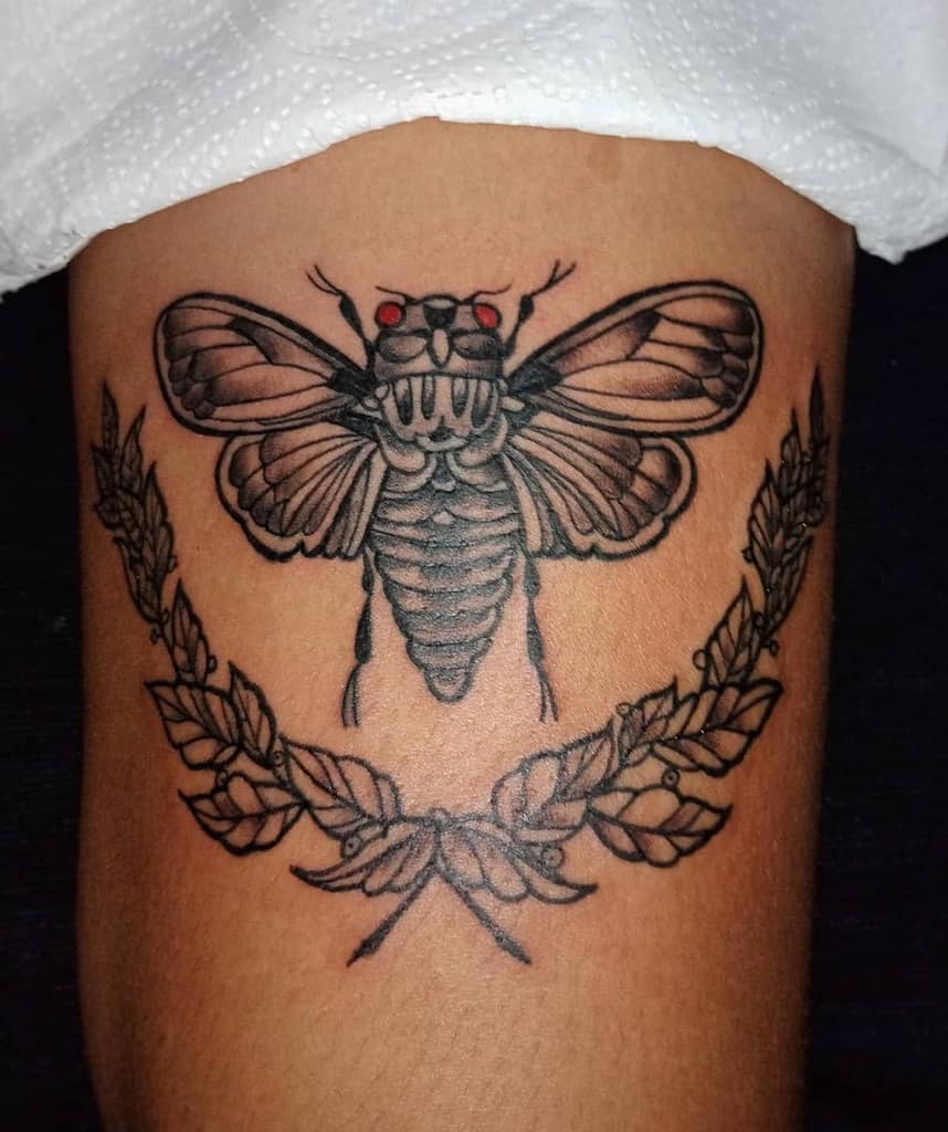Tattoo of a cicada with red eyes and wings above a laurel wreath on a thigh