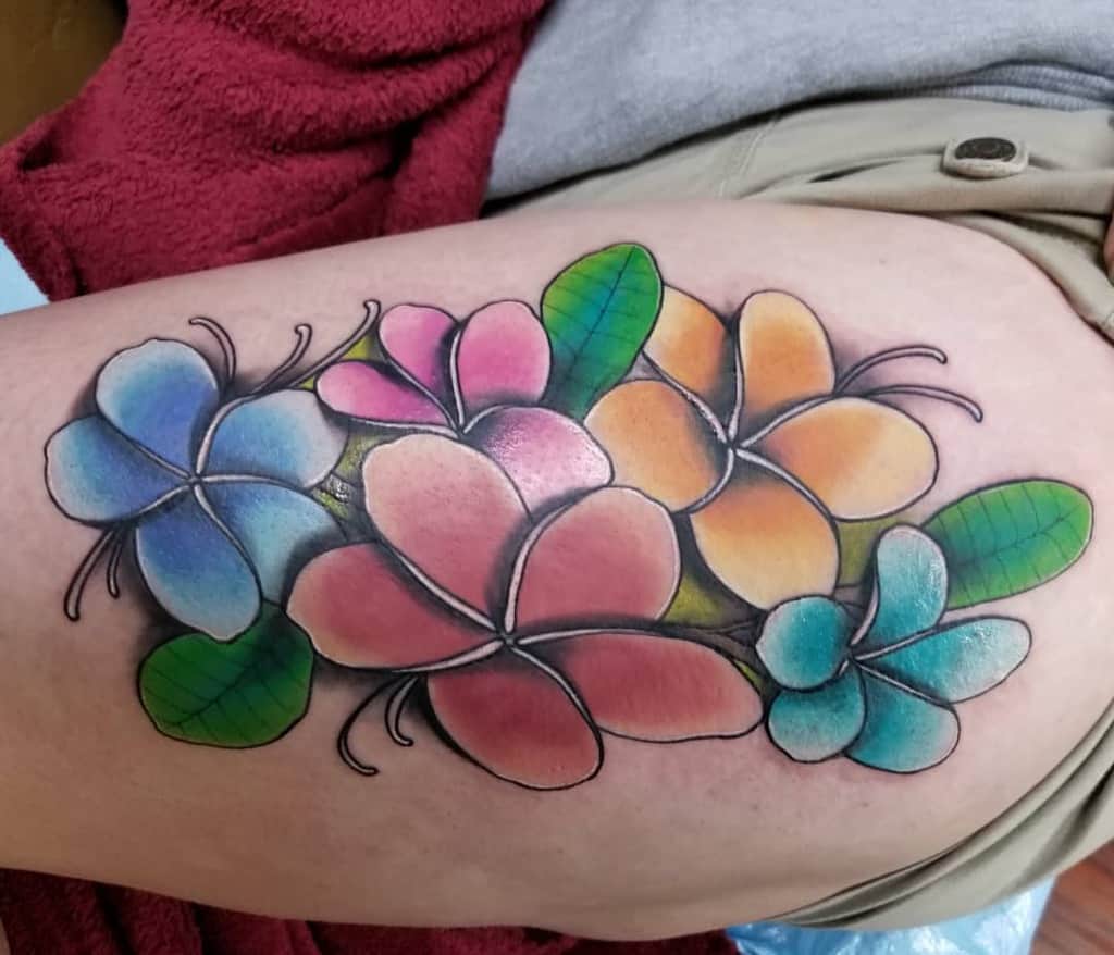 A vibrant plumeria tattoo adorns the skin, boasting pink, blue, and orange petals alongside lush green leaves