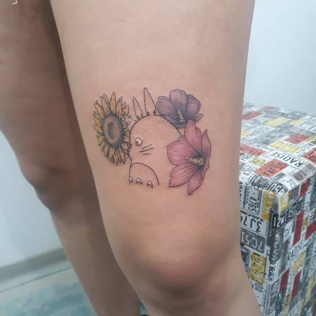 Minimalist Totoro thigh tattoo surrounded by a sunflower and pink flower, creating a delicate and nature-inspired look
