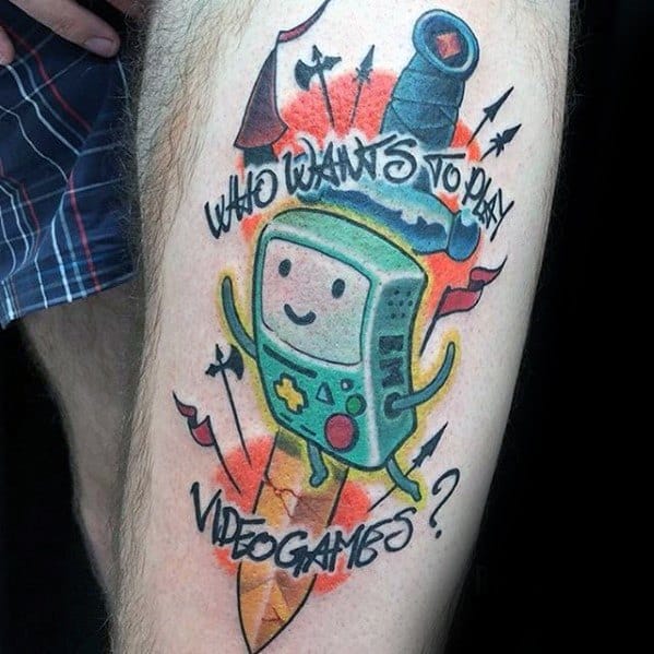 Adventure Time tattoo of BMO with the quote “Who wants to play video games?” surrounded by arrows and vibrant color splashes, inked on the thigh.