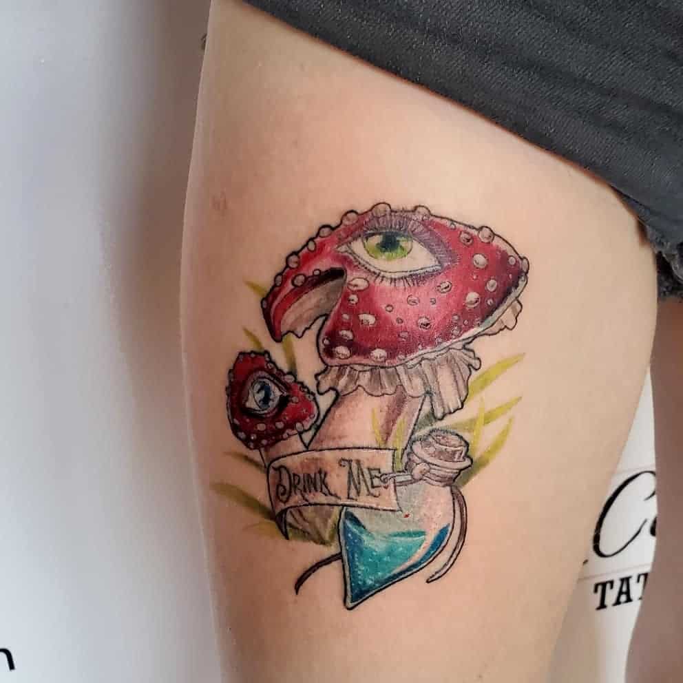 A colorful tattoo of a mushroom with an eye, labeled "Drink Me" on a ribbon, inspired by Alice in Wonderland