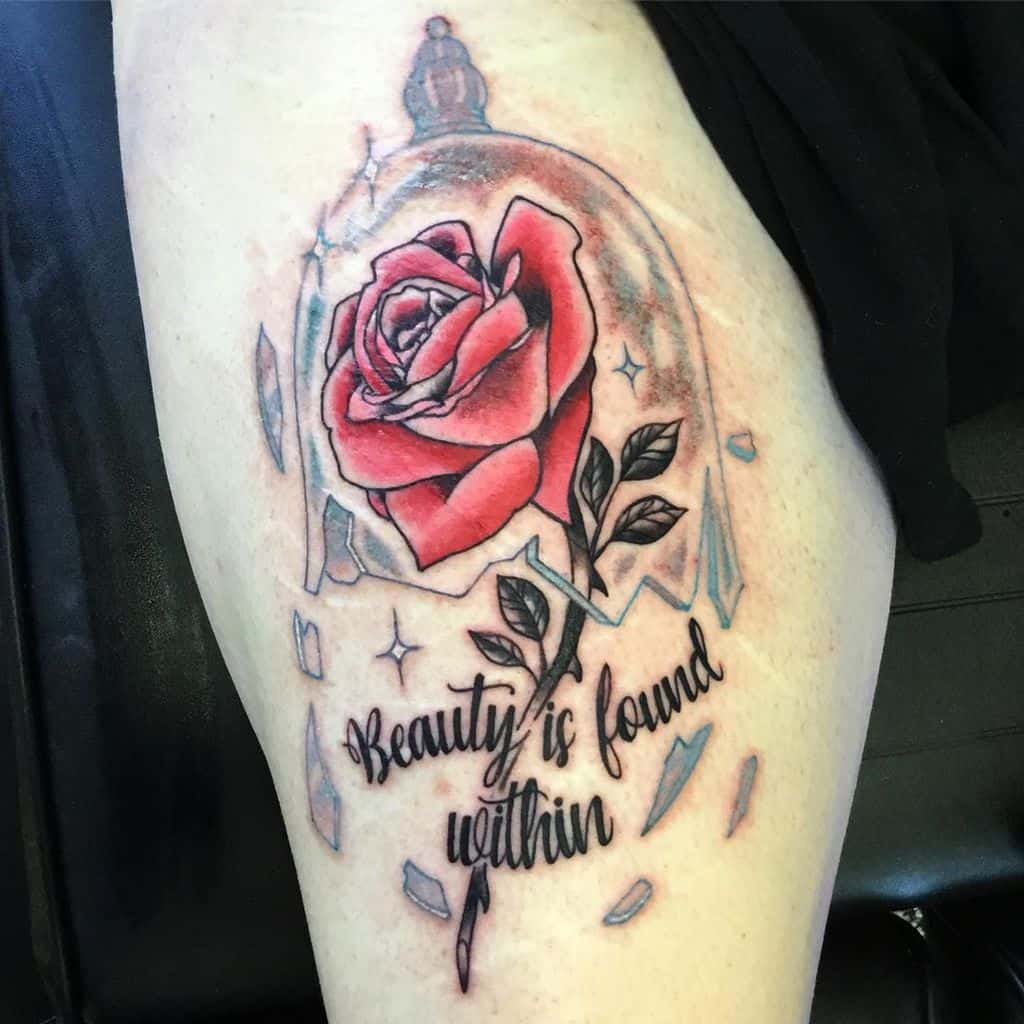 Tattoo of a red rose under glass with shards and "Beauty is found within" text on a thigh