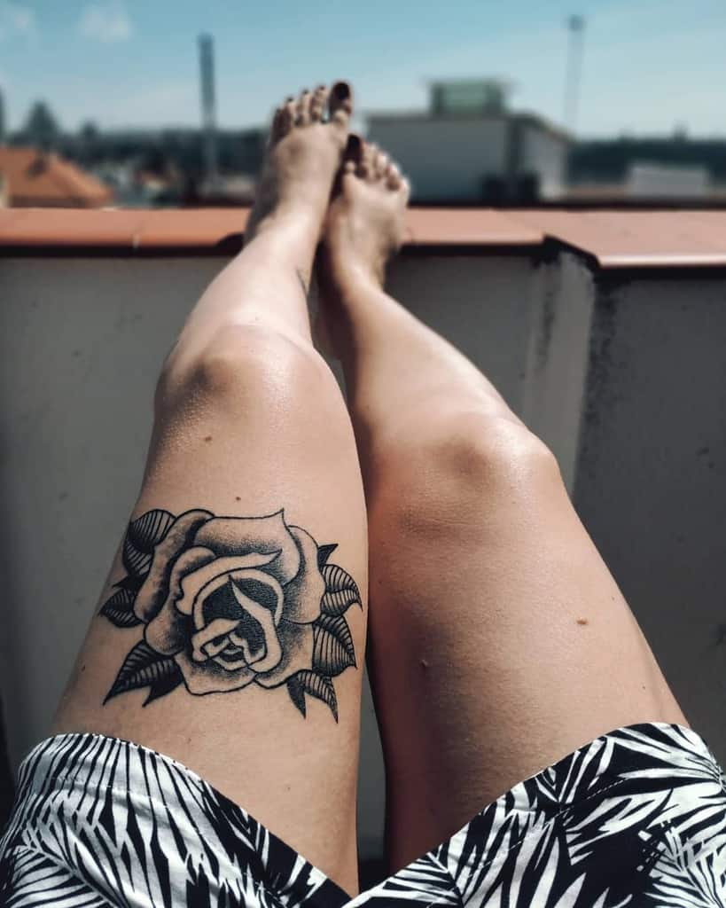 A black and white tattoo of a detailed rose with shading, placed on the thigh, surrounded by intricate leaves for added contrast