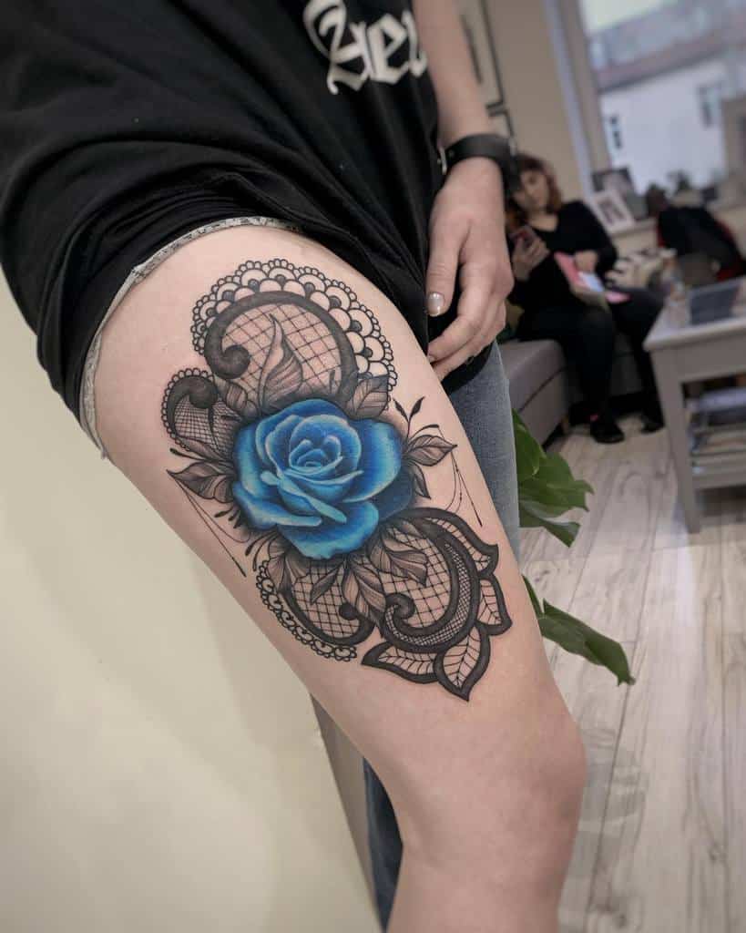 A stunning blue rose tattoo on the thigh, surrounded by intricate black lace patterns and shading for a detailed, elegant design