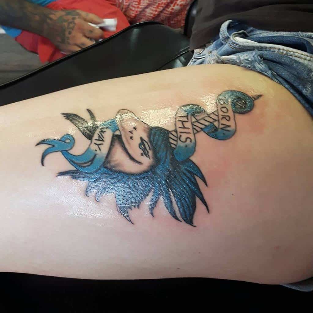Thigh tattoo featuring a blue-haired figure with a 'Born This Way' ribbon wrapped around, blending bold colors and artistic detail