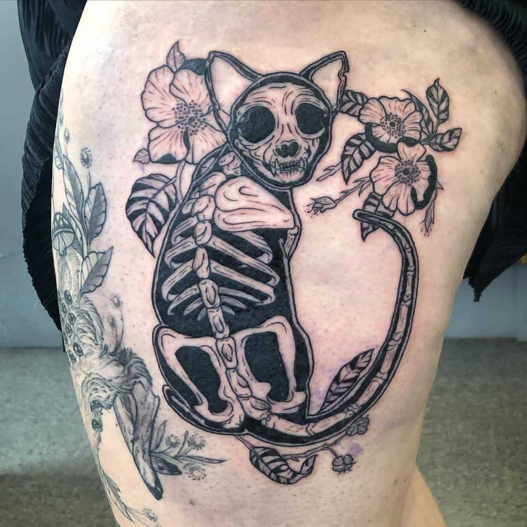 Tattoo of a cat skull and skeleton adorned with flowers on an upper thigh