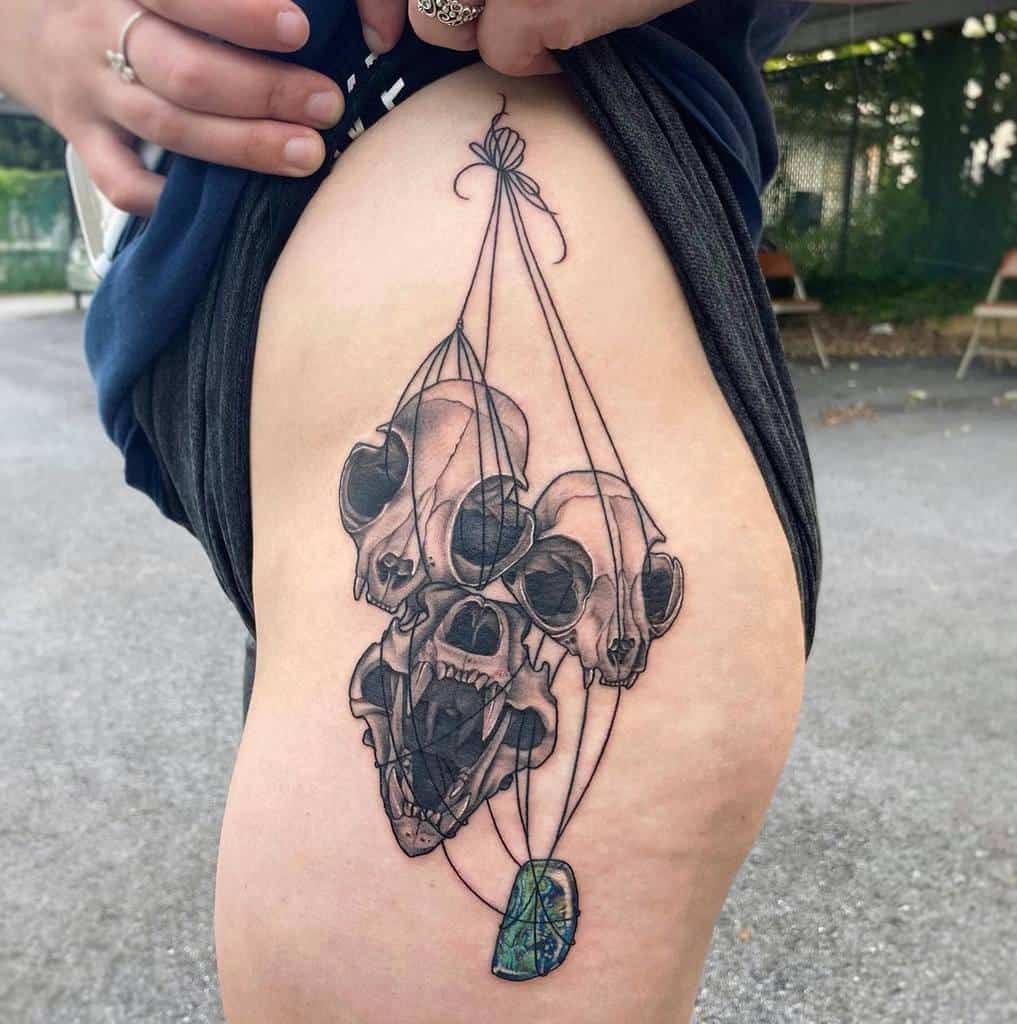 A tattoo of three detailed cat skulls tied together with strings, hanging from a stone pendant on the thigh