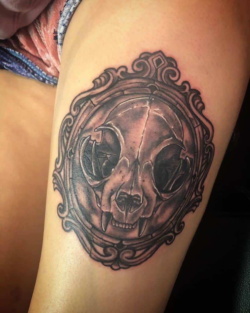 Tattoo of a cat skull within an ornate circular frame on an upper thigh