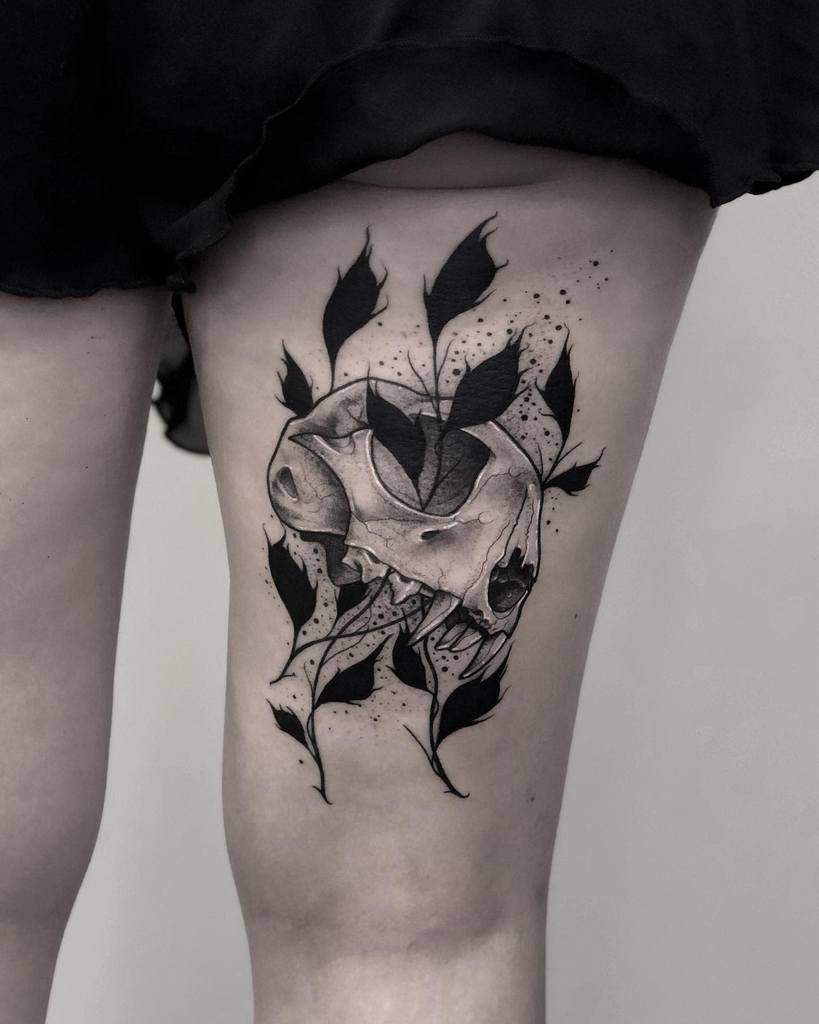 Thigh tattoo of a cat skull surrounded by black leaves and intricate decorative elements