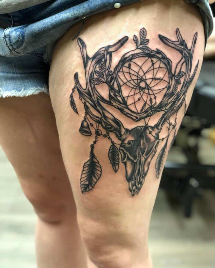 Tattoo of a deer skull with antlers, dreamcatcher, and feathers beautifully entwined on a person's upper thigh