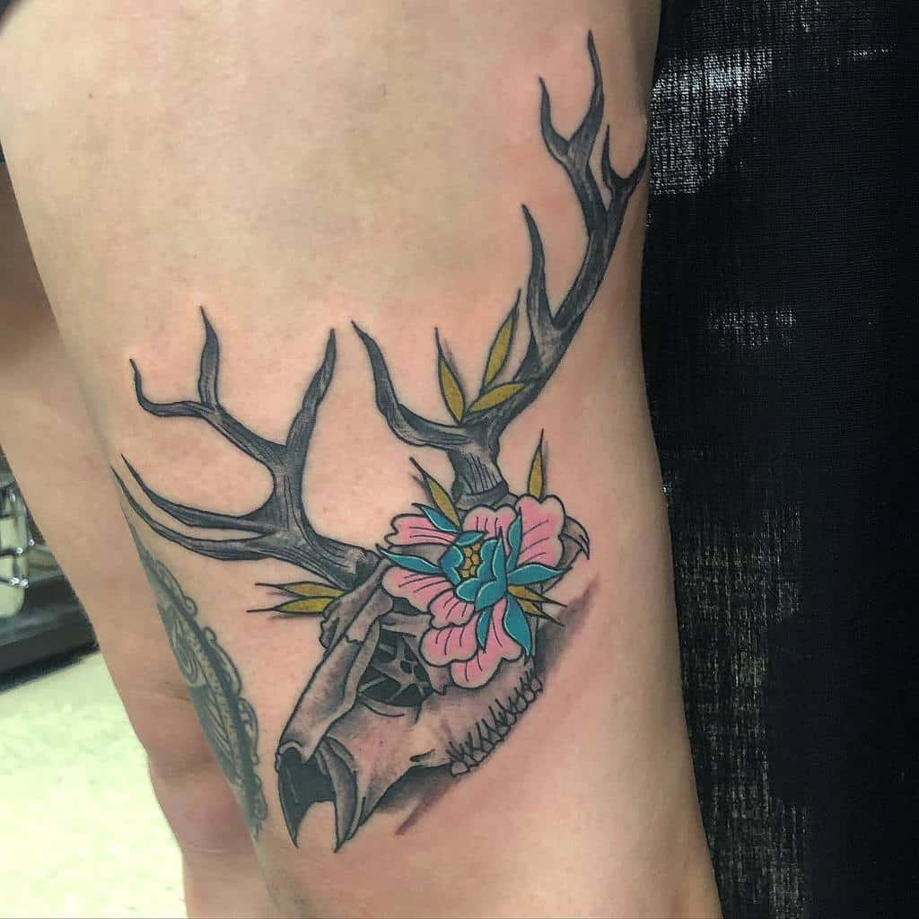 Tattoo of a deer skull with antlers, adorned with a blue and pink flower and green leaves