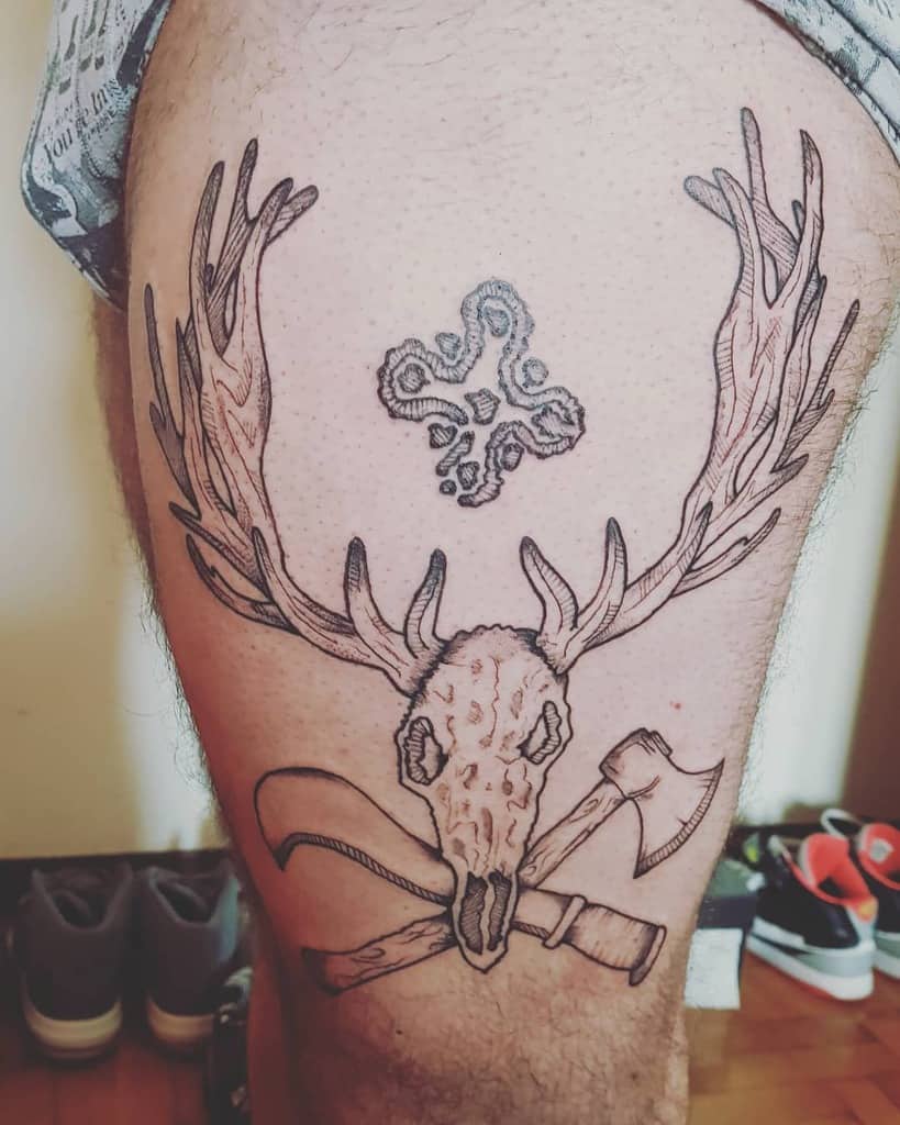 Deer skull tattoo with antlers, featuring an ax and a cross design on a thigh