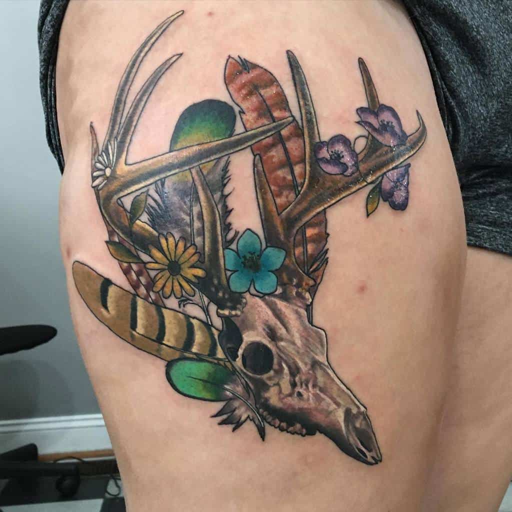 Tattoo of a deer skull with antlers adorned with feathers and flowers beautifully inked on a person's thigh