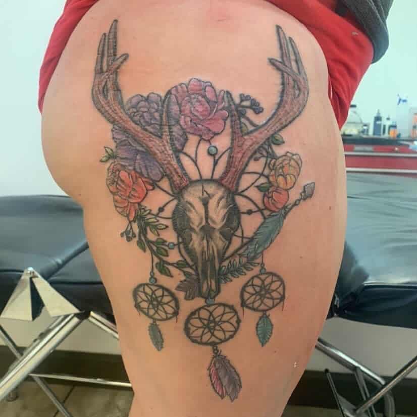 Tattoo of a deer skull adorned with dreamcatchers and flowers on the thigh
