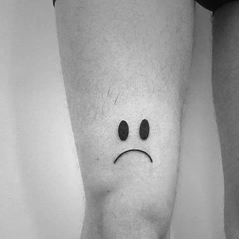 Sad face emoji tattoo on the front of a person's upper leg in black and white
