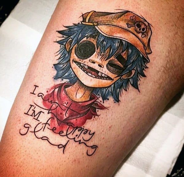 Color tattoo of 2D from Gorillaz with a wide grin, blue hair, skull cap, and the lyrics “I am happy, I’m feeling glad” beneath in stylized text.