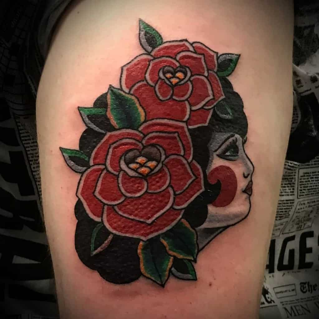 A bold tattoo of a woman with two large red roses in her hair, with traditional tattoo style outlines, on the thigh