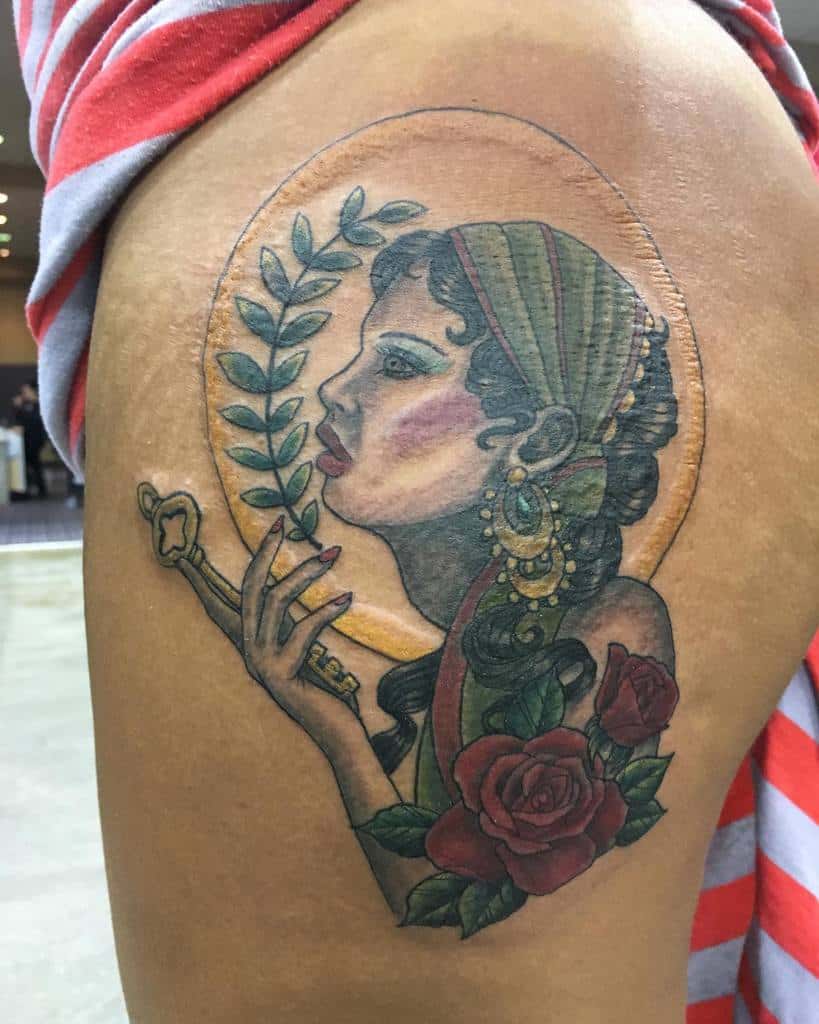A vibrant tattoo of a gypsy woman with a key and rose, surrounded by a leafy wreath, on the thigh