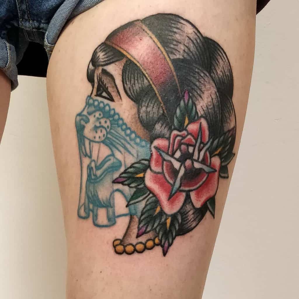 A colorful tattoo of a gypsy woman with a veil and a rose in her hair, alongside a skull and floral elements, on the thigh