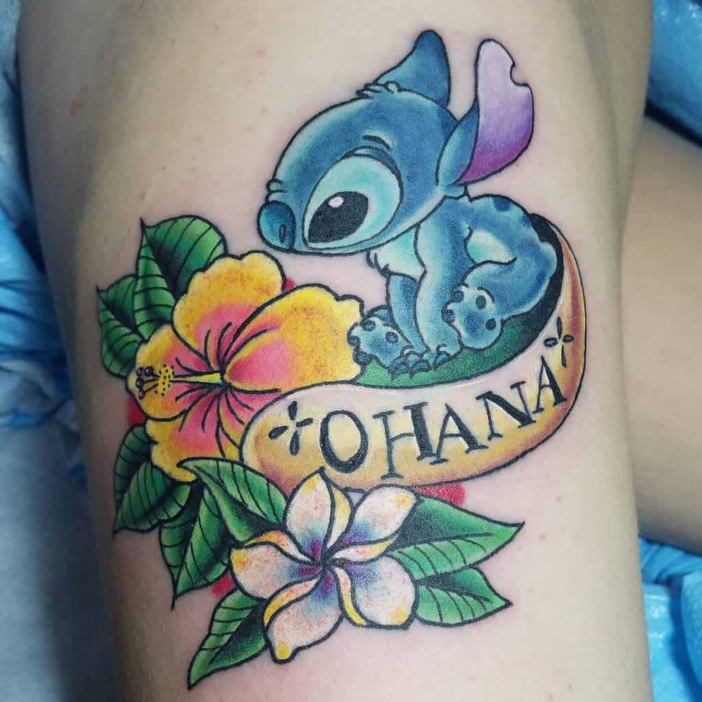 Thigh tattoo of a blue cartoon creature sitting with flowers and a banner that reads "OHANA