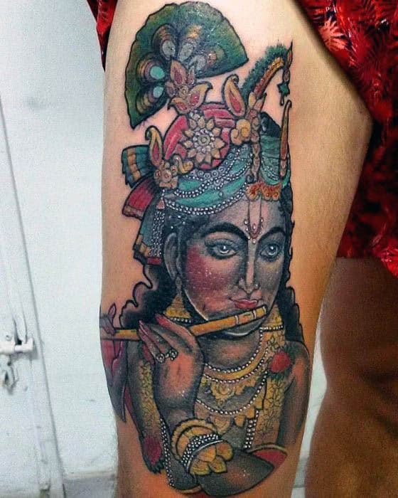 Colorful Krishna tattoo on the thigh showing him playing the flute, adorned with ornate jewelry, a vibrant crown, and a detailed peacock feather above.