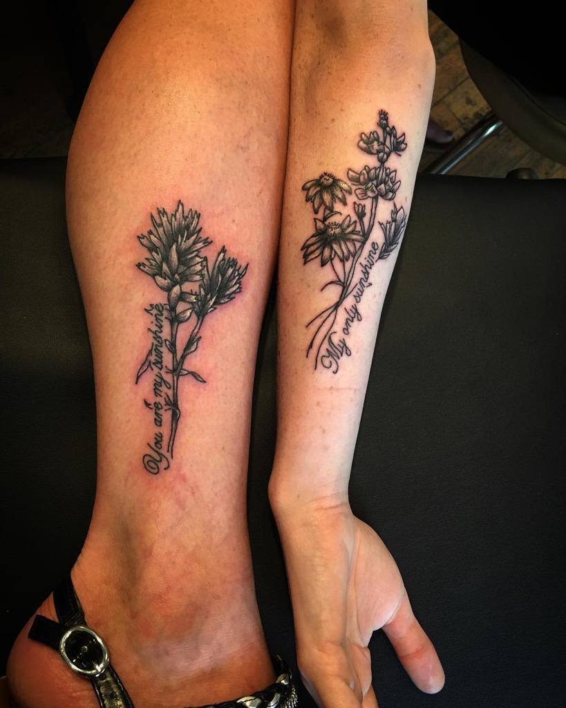Matching tattoos featuring wildflowers and script reading 'You are my sunshine' and 'My only sunshine,' delicately placed on the leg and forearm